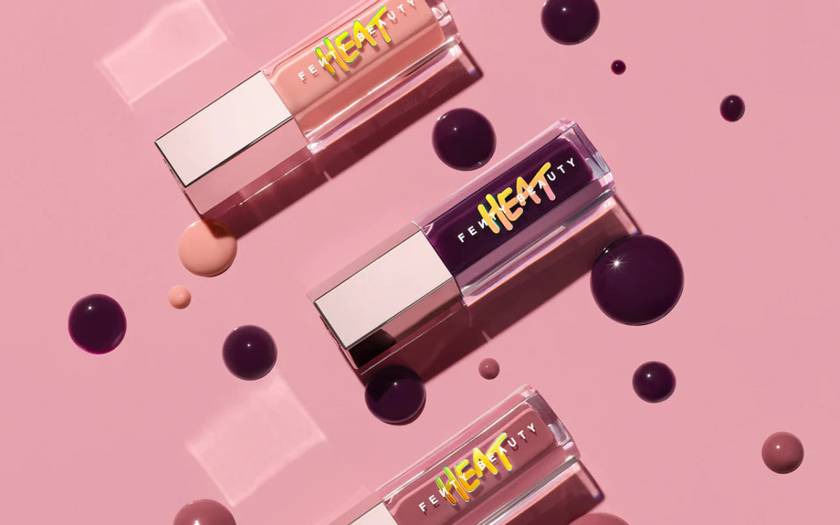 Three Fenty Beauty Heat liquid lip products arranged diagonally on a pink background with glossy liquid droplets. The products have clear rectangular tubes showing pink, nude, and mauve shades, with metallic silver caps and yellow 'HEAT' branding.